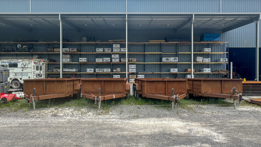 Wide-angle image capturing the full profile of an unpainted MSA dump trailer on a gravel lot.